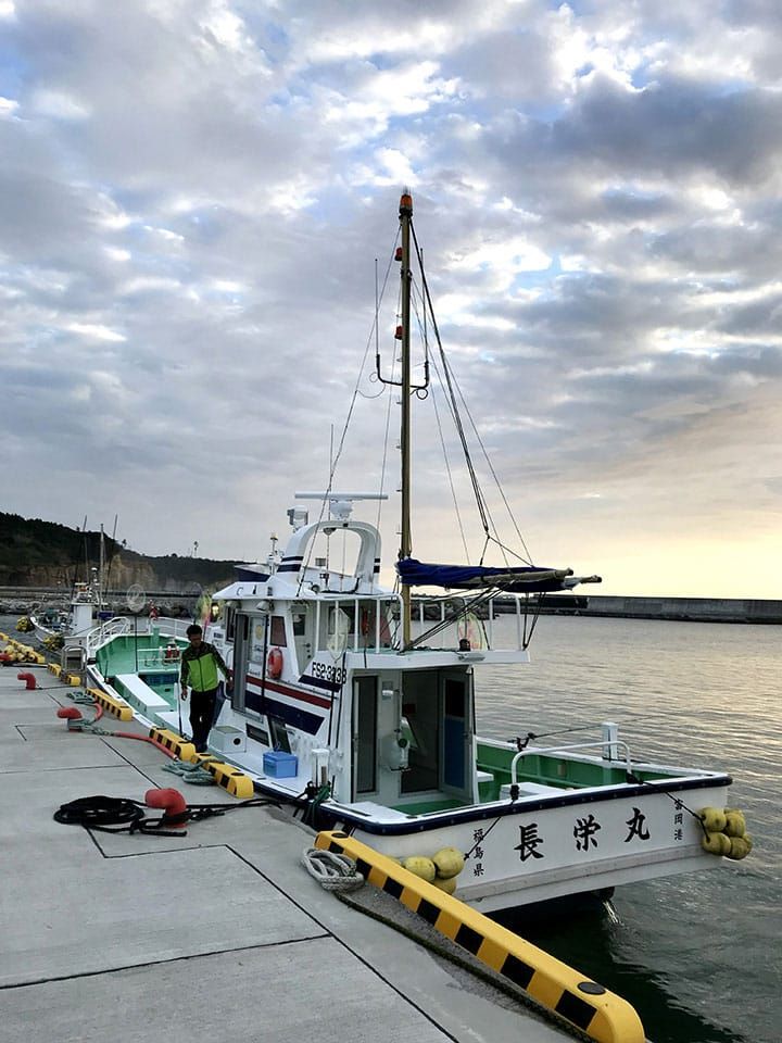 朝焼けの空のもと、富岡漁港の岸壁に停泊している遊漁船「長栄丸」の上を男性の漁師が歩いている様子