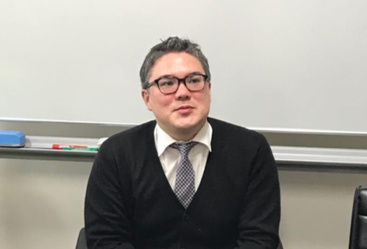 カナダ人のマクマイケル・ウィリアムさんがホワイトボードの前でインタビューに応じている様子の社員