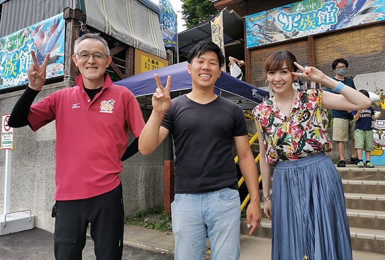 職員の男性と鰐さん、女性インタビュアーが入場口で笑顔でピースのポーズをとっている集合写真