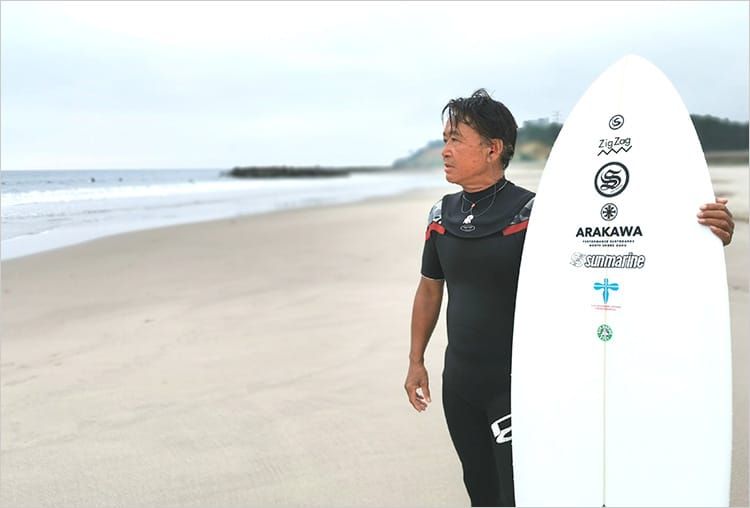 北浜海岸の砂浜で、ウエットスーツを身に着けた鈴木さんが、サーフボードを片手に海の向こうを見つめている写真