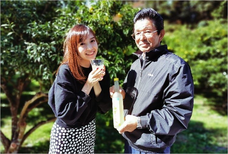 ゆずの果樹園を背景に、高橋麻里江さんと松本広行さんが「ゆず里愛」の瓶とクラスを片手に笑みを浮かべている写真。
