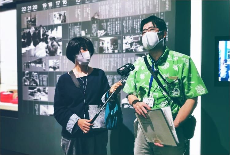 展示パネルの前で女性インタビュアーからの質問に応じる、語り部の泉田淳さん