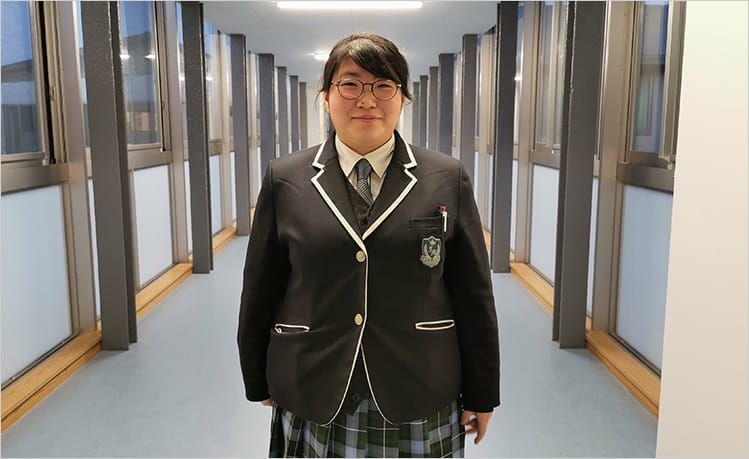 学校の廊下に立ち、制服姿で笑みを浮かべる畠山歩さんの写真