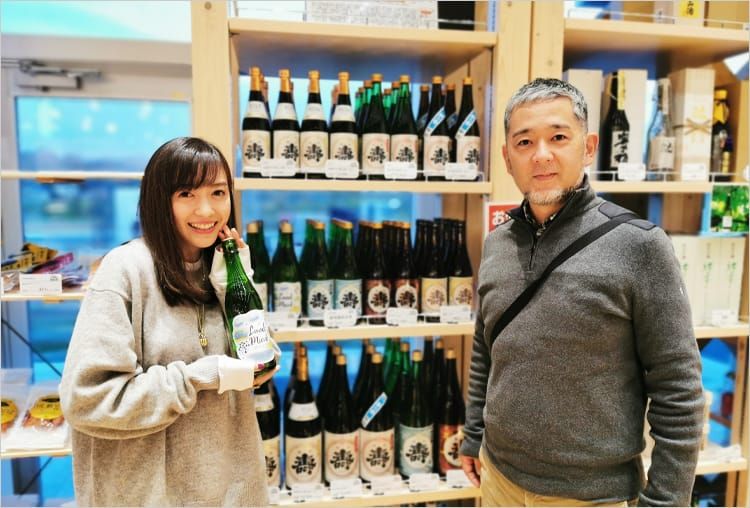 道の駅なみえの店内の棚を背景に、鈴木大介さんとボトルを持った高橋麻里江さんが笑顔で並んでいる写真