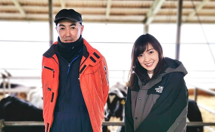 牛舎を背景に田中一正さんと高橋万里恵さんが笑顔で並んでいる写真
