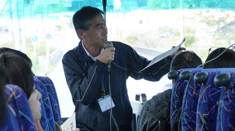 移動中のバスの車内で安藤宏さんがマイクを持ち、窓の外を見ながら乗客に説明を行っている様子