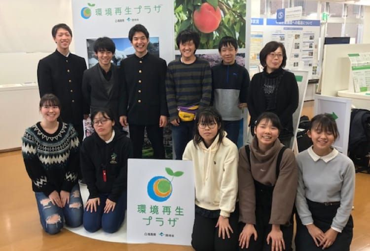 環境再生プラザの展示パネルを背景に、参加した教諭と十名の学生らが笑顔で二列に並んでいる集合写真